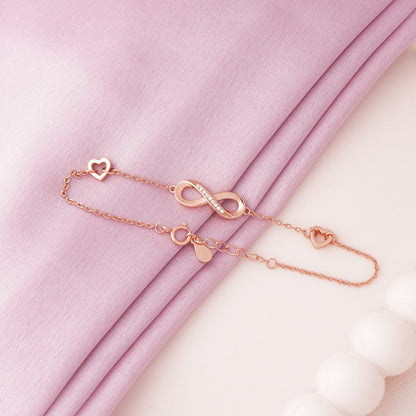 Rose gold bracelet with heart, infinity, and circle charms on a pink fabric background